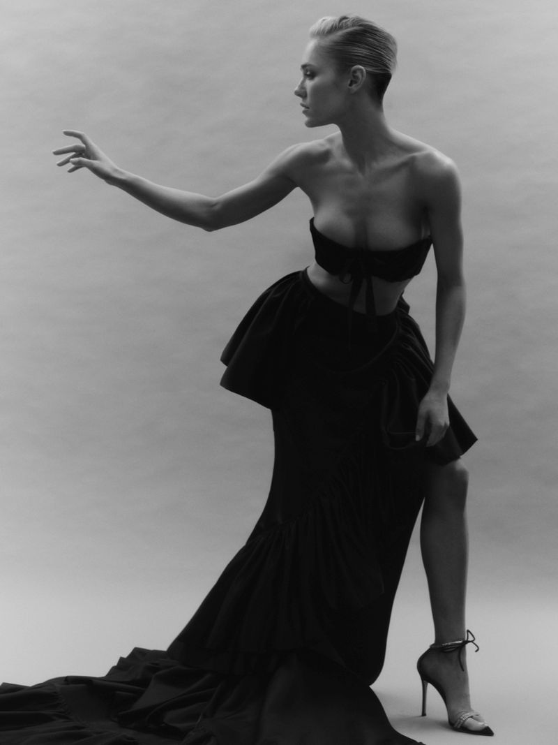 Monochrome studio shot of a stylish adult woman in a dark top and a flowing skirt, striking a pose against a neutral background.