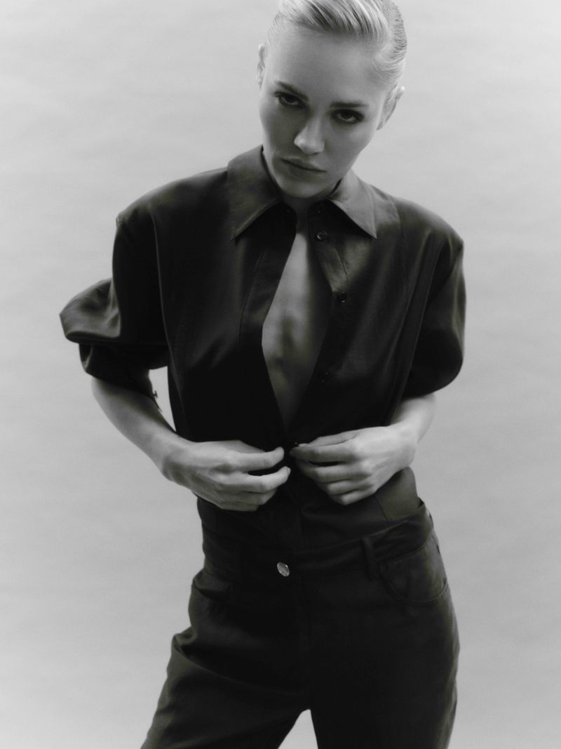 Black and white portrait of a blonde adult female fashion model buttoning a black collared shirt and high-waisted pants, posing indoors.