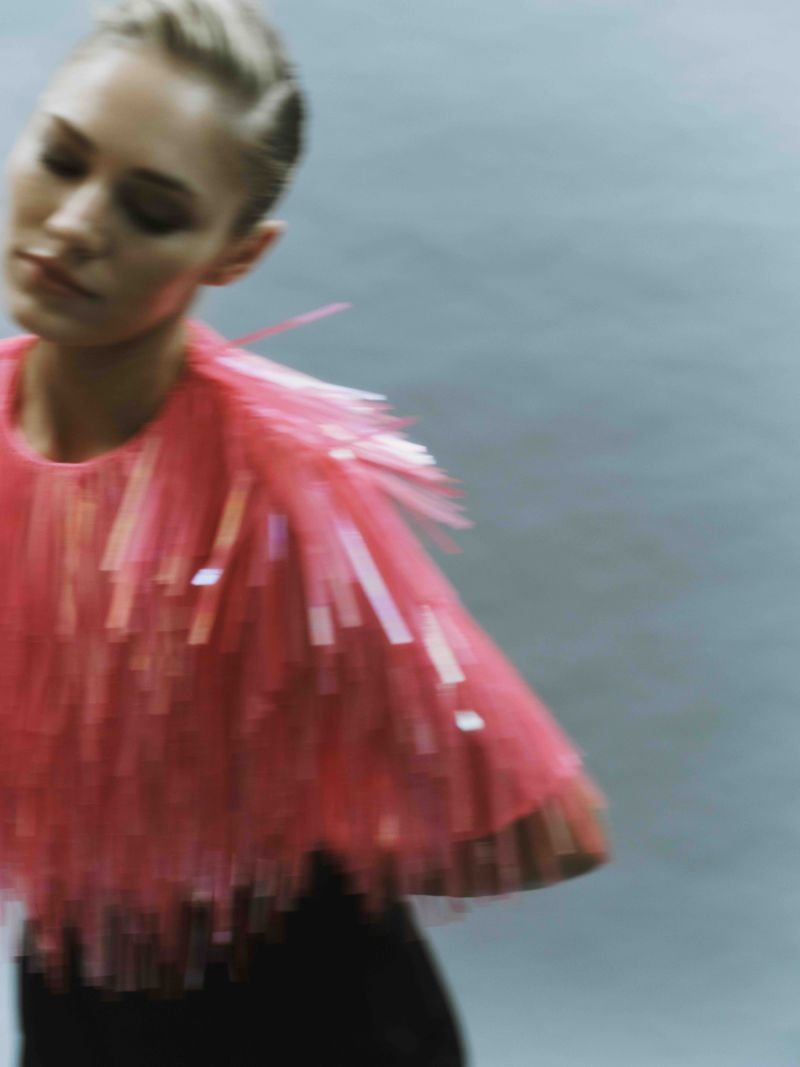 Woman wearing a neon pink fringe top in a blurred motion fashion shot against a grey backdrop in a studio with soft, diffused lighting.