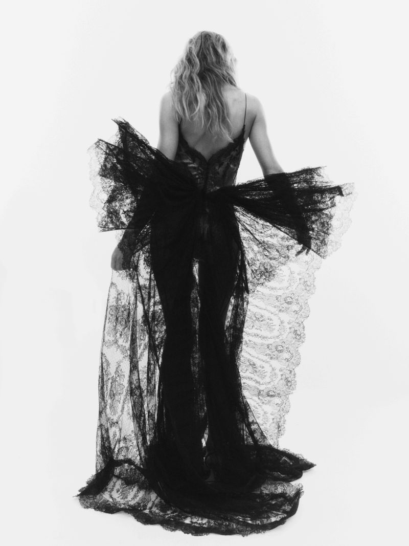Back view of a woman in a black lace dress with a large bow, posing against a white background, showcasing fashion and elegance.