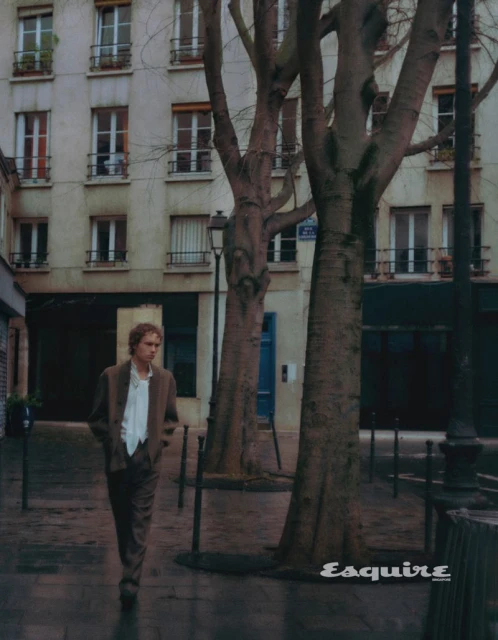 Man in brown suit walking on a wet city street lined with trees and buildings in an editorial fashion shot during the daytime.