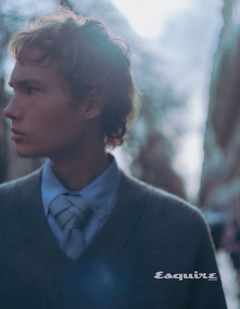 Portrait of a young adult male with fair skin and curly hair wearing a sweater and collared shirt against a blurred background. 