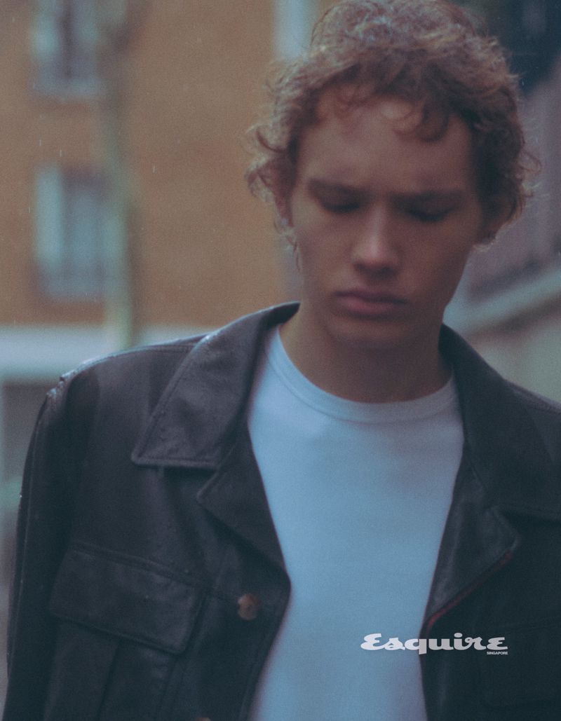 Young man in a black leather jacket stands outside on a city street, with rain drops in the air, and the Esquire logo in the corner.