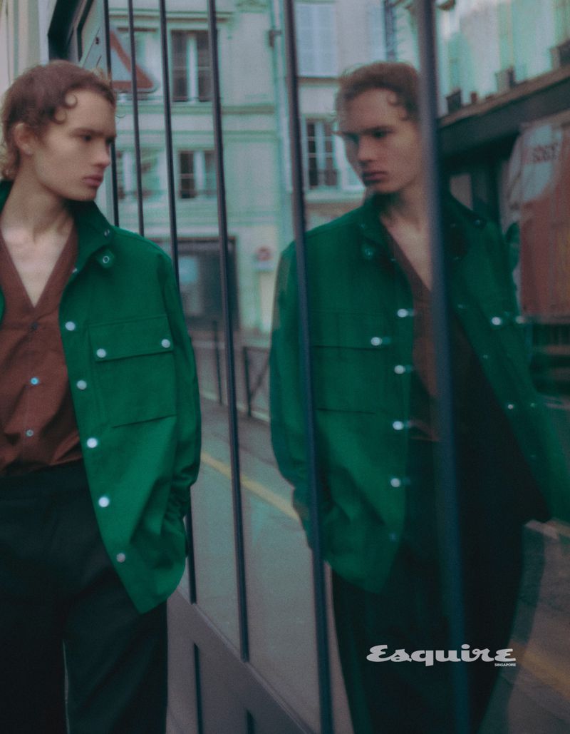 Male fashion model poses in a green jacket and brown shirt with an urban setting reflected in a nearby glass surface.