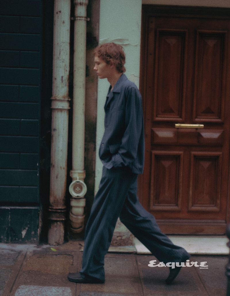 A slender man in a blue pajama-style suit strides along a city sidewalk past a blue wall and a brown door in a fashion editorial.
