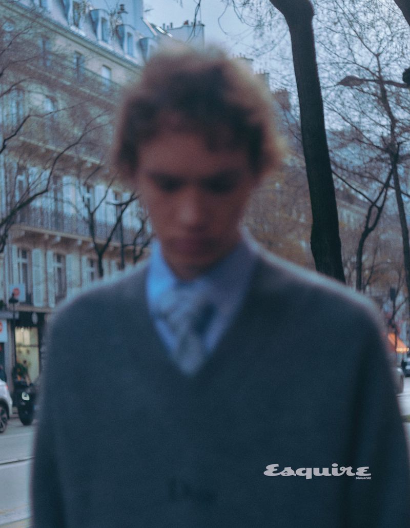 Blurred portrait of a young man in a grey v-neck sweater and blue shirt walking outside in a European city with vintage color grading.