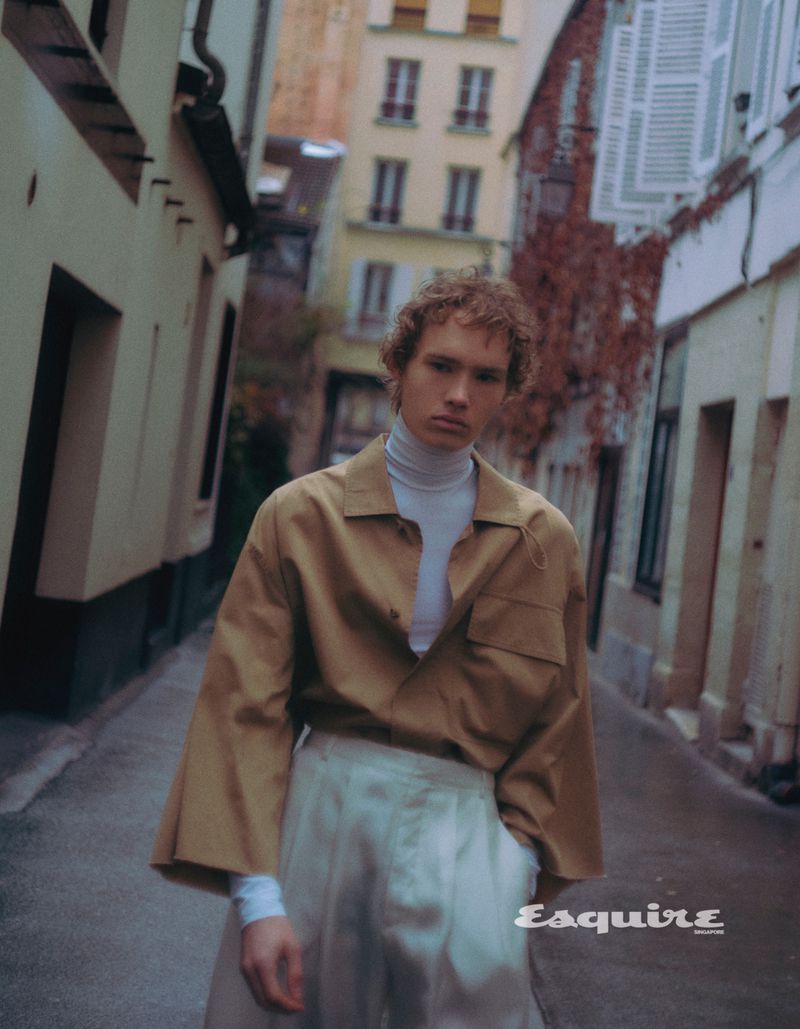 Man in tan shirt, white turtleneck, and wide-leg pants stands on a narrow street lined with tall buildings and a European vibe.