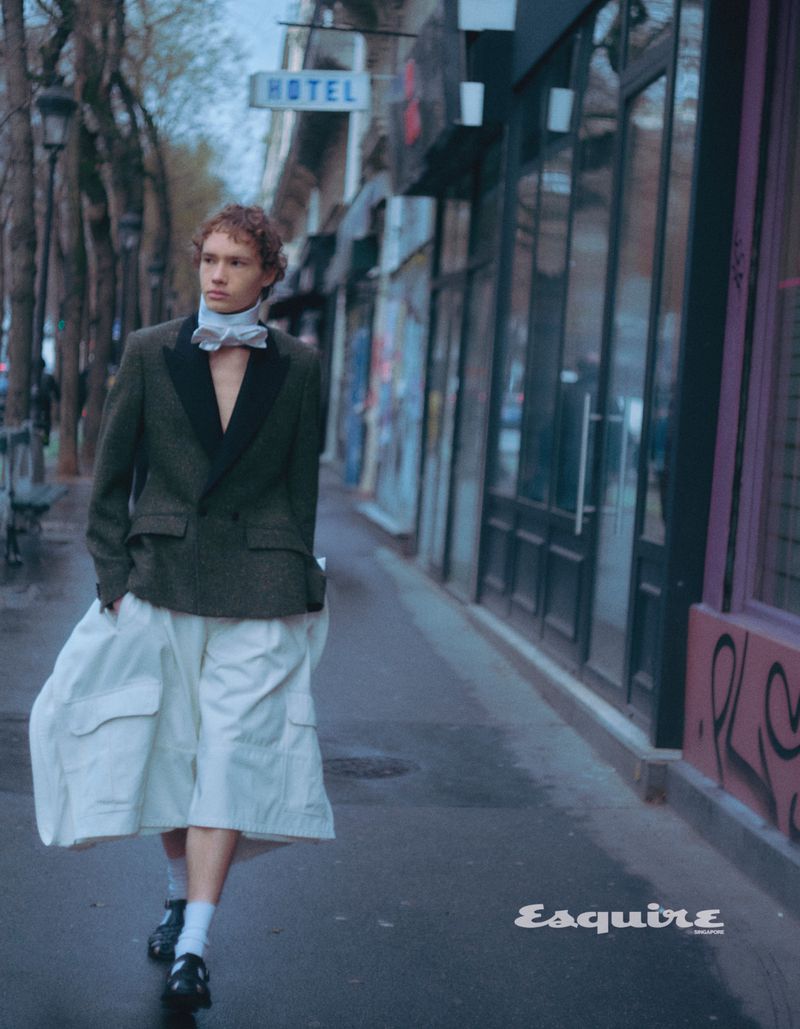 A young man walks on a city street wearing a suit jacket, large white shorts, socks, and shoes. Trees and buildings line the street.