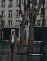 Model walking down a wet urban street past large trees in front of a multi-story building facade.
