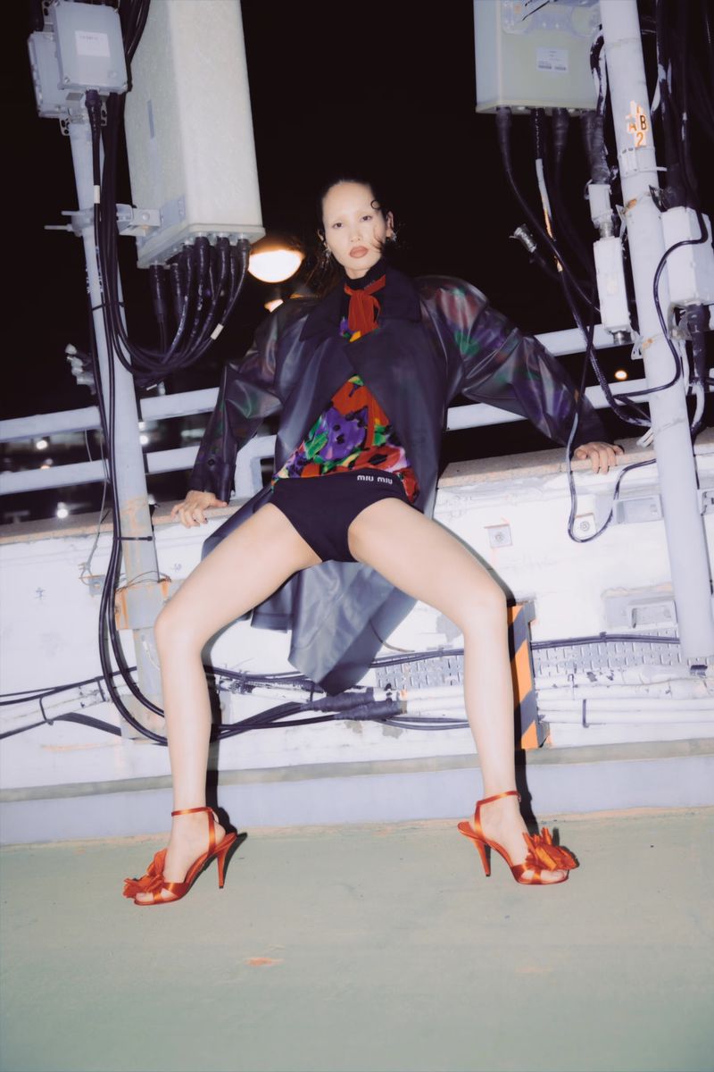 A stylish young woman poses on a rooftop in a floral shirt, black shorts, and red heels next to utility boxes, shot at night.