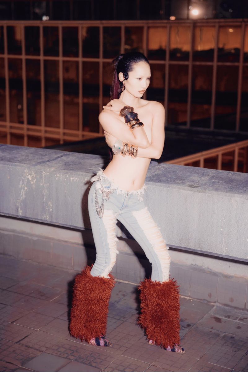 A woman poses on a rooftop in a fashion shot featuring distressed denim, fur boots, and dramatic arm jewelry against an urban backdrop.