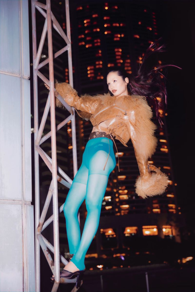 An Asian woman strikes a pose on metal framework; dressed in a tan jacket, teal bottoms, and black heels in front of a city skyline.