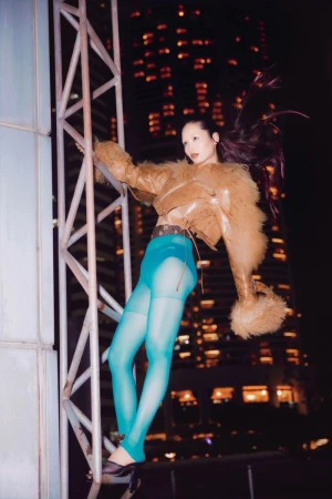 An Asian woman strikes a pose on metal framework; dressed in a tan jacket, teal bottoms, and black heels in front of a city skyline.