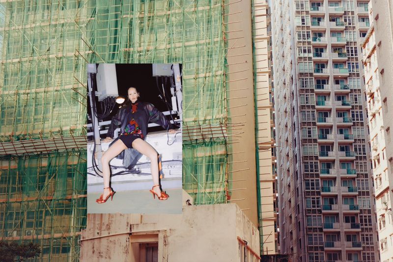A woman poses in high heels on a building, with a construction site and high-rise apartments in the background, creating an urban scene.