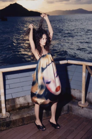 Woman in unique face-print dress strikes pose against ocean backdrop. Dark hair, evening light, concrete deck with railing.