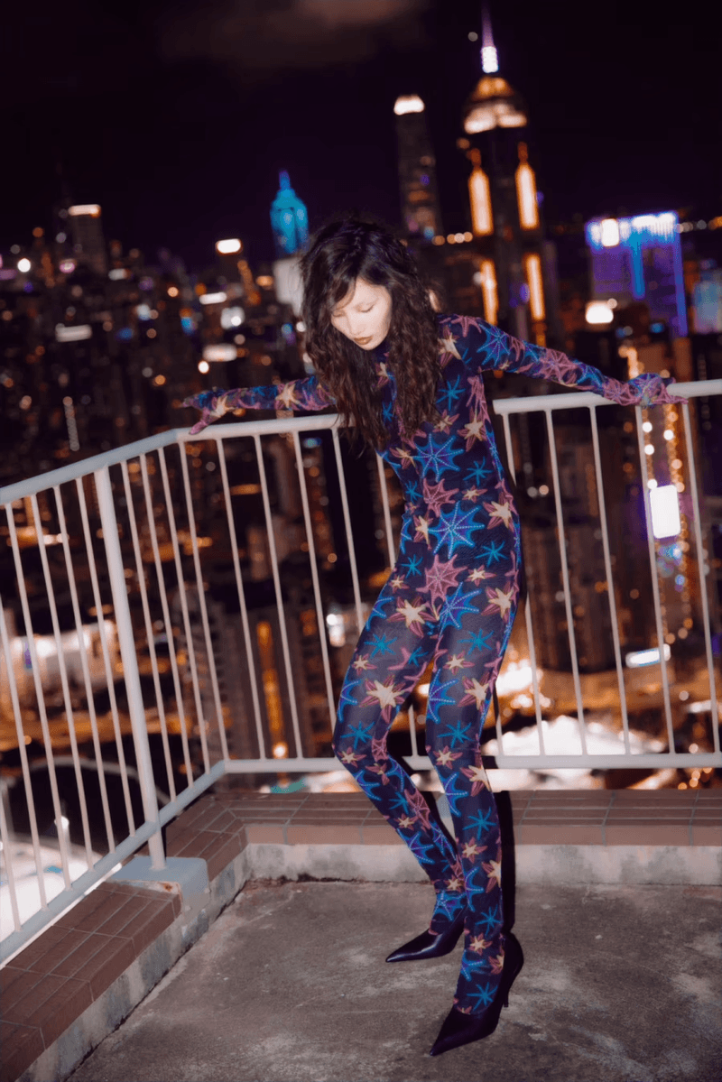 Woman in a patterned bodysuit poses on a rooftop at night, with a cityscape of illuminated buildings in the background.