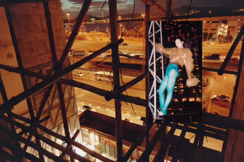 Fashion photo of an adult female model wearing a brown jacket and blue tights, posing on scaffolding with an urban city view at night.