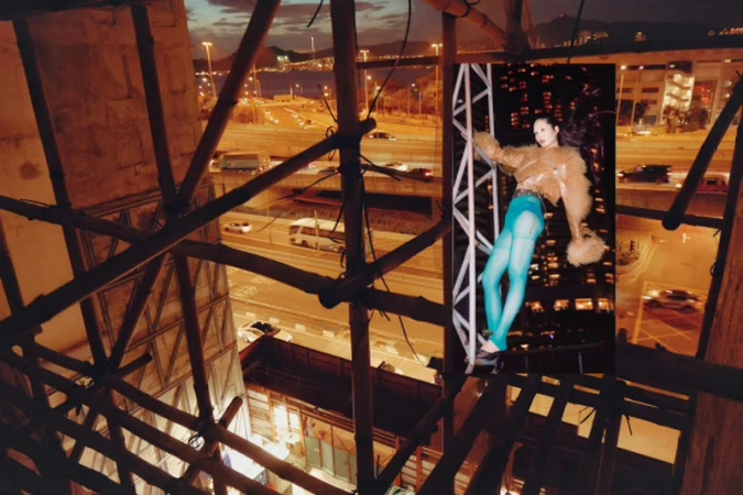 Fashion photo of an adult female model wearing a brown jacket and blue tights, posing on scaffolding with an urban city view at night.