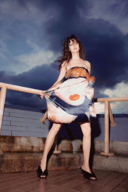 A woman stands on a rooftop at night, wearing a strapless dress with an abstract pattern, black heels, and with a dramatic, cloudy sky.