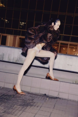 Fashion shot of a woman in a fur coat climbing steps outside at night, wearing pantyhose and heels, with a city building background.