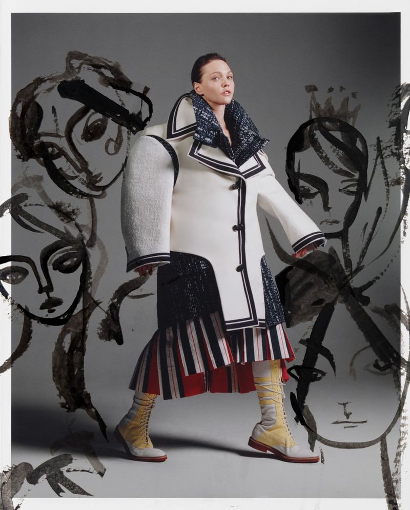 Fashion editorial shot: Woman in a white coat with black trim poses amid abstract face drawings, showcasing avant-garde style and design.