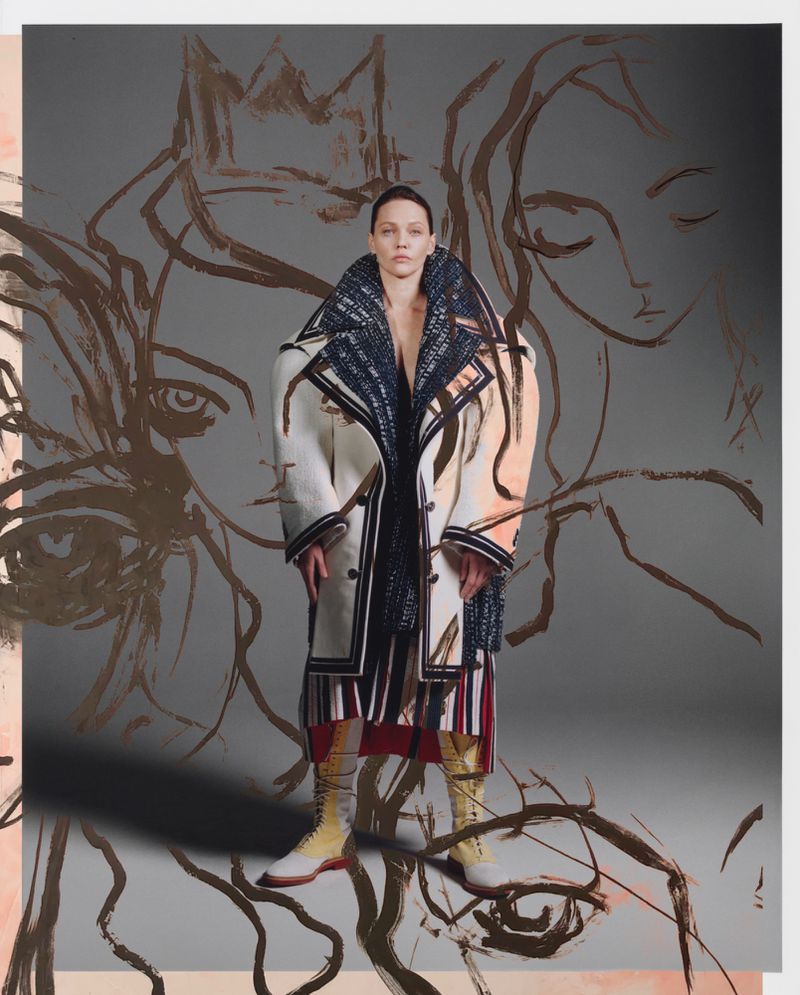 Fashion shot: adult female model poses in an elaborate coat with a sketched face in the background, creating an artful and high-fashion image.