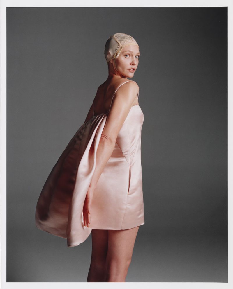 Blonde woman in a pale pink dress and cap looks over her shoulder in a studio shot against a gray background, fashion portrait.