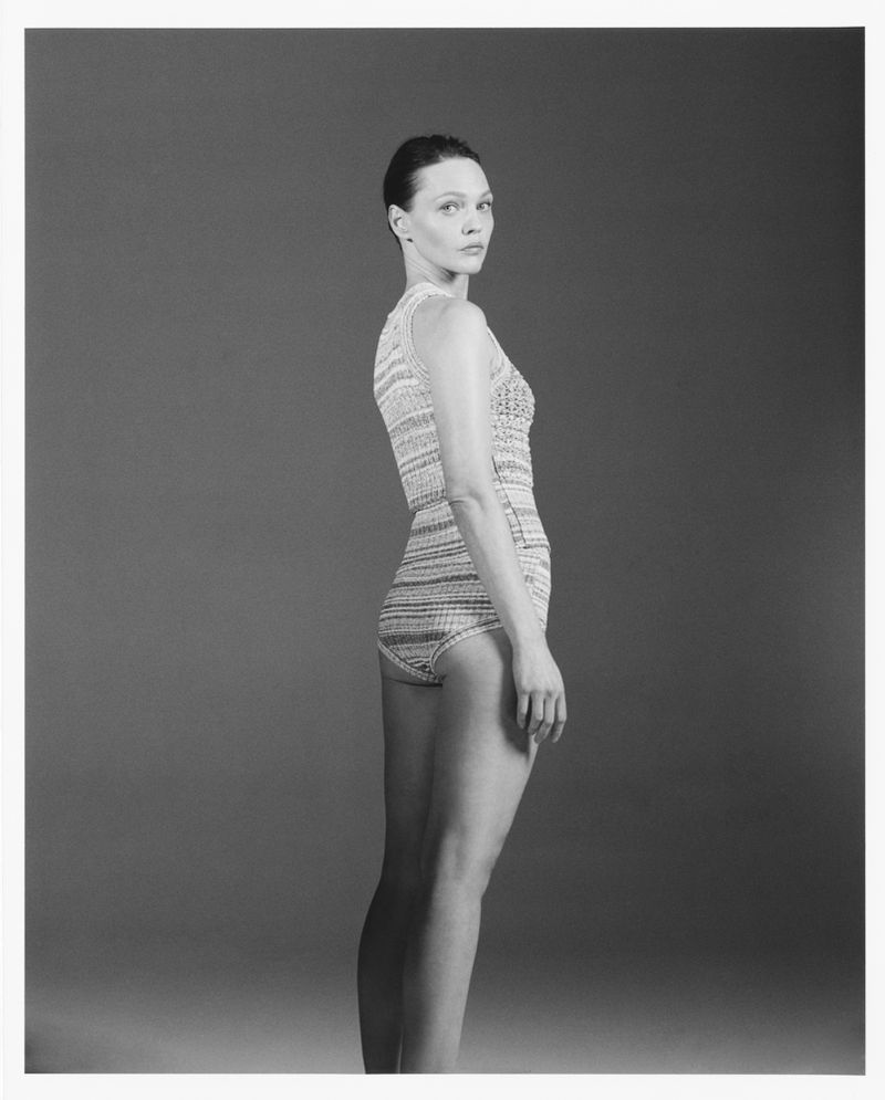 Black and white studio portrait of a young adult woman wearing a knit bodysuit, turned slightly to the side with an intense gaze.