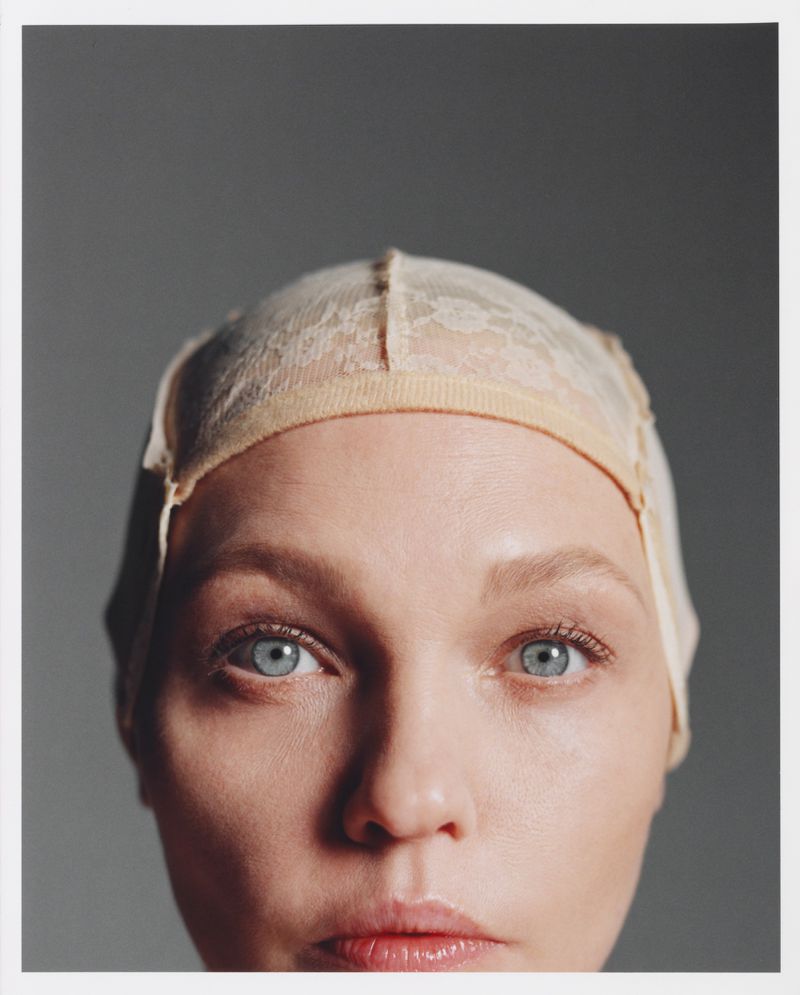 Close-up portrait of a woman with fair skin and blue eyes wearing a beige lace wig cap against a solid grey backdrop in a studio setting.