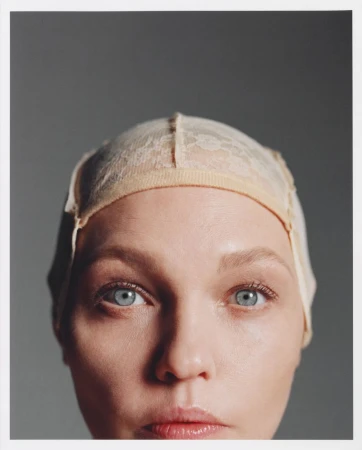 Close-up portrait of a woman with fair skin and blue eyes wearing a beige lace wig cap against a solid grey backdrop in a studio setting.