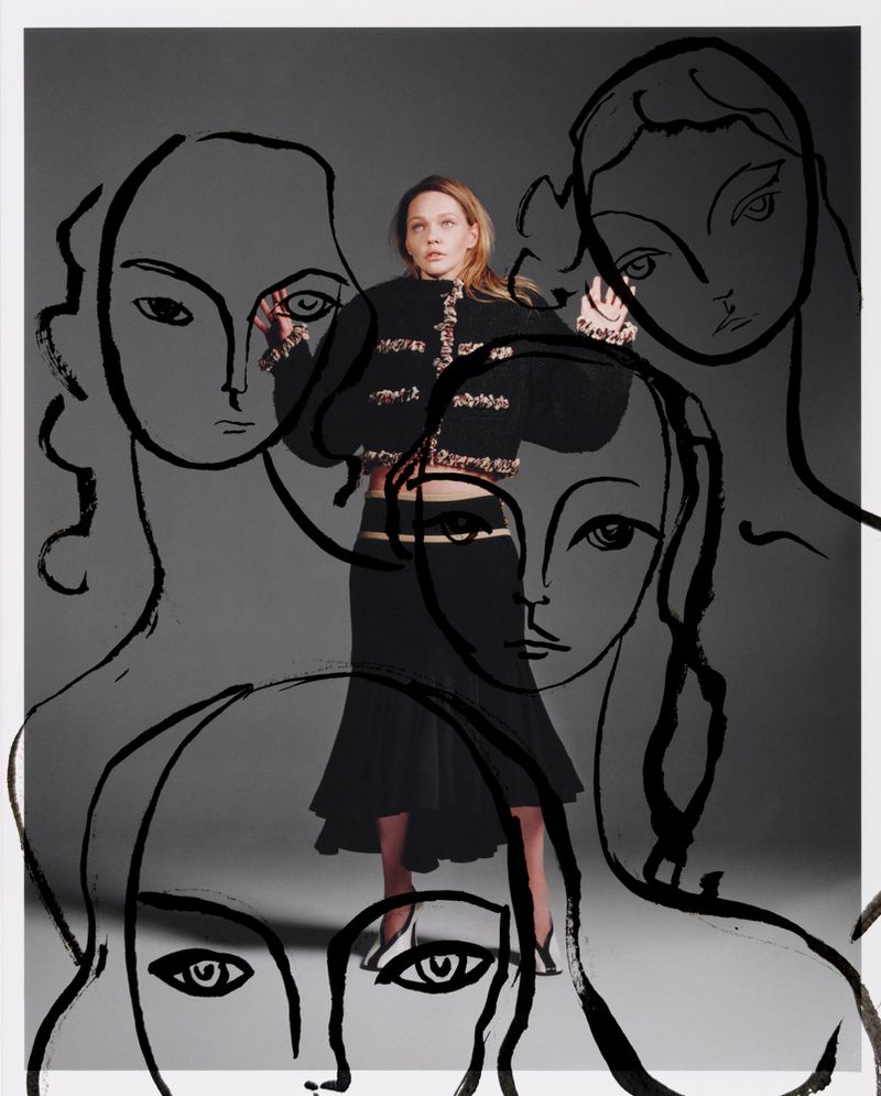 Woman in a black skirt and jacket poses in front of sketched faces against a gray background in a studio fashion shot.