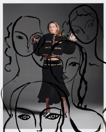 Woman in a black skirt and jacket poses in front of sketched faces against a gray background in a studio fashion shot.