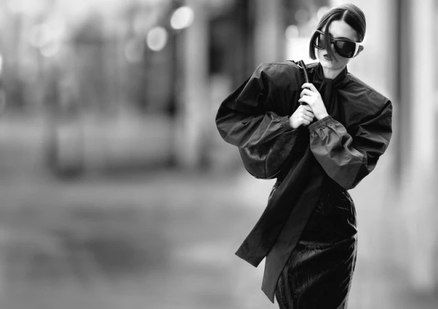 Fashion portrait of a woman in sunglasses wearing a shiny black outfit against an out-of-focus urban background in black and white.