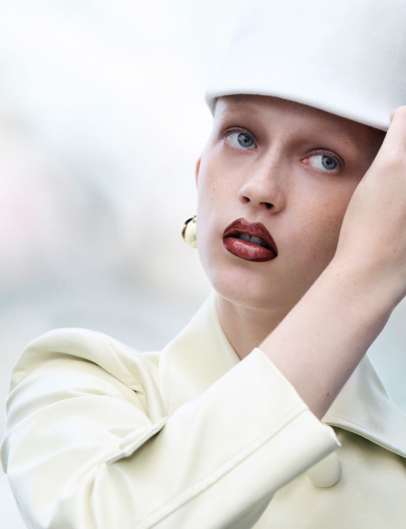 Close-up fashion portrait of a woman in a cream coat and white hat, with dark lipstick and gold earrings against a blurred background.