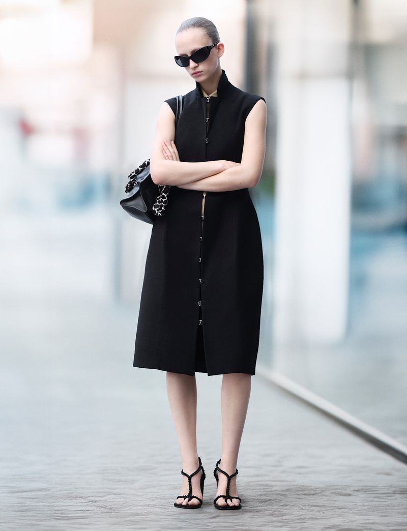 Fashionable woman in black dress, sunglasses, and strappy heels stands with crossed arms in an urban setting, showcasing modern style.