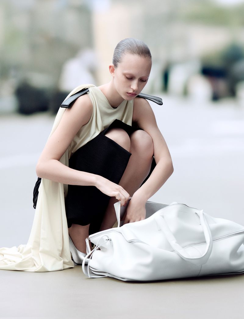A slender woman kneels, her gaze fixed on a large white bag, her minimalist outfit and slicked-back hair accentuating her modern look.