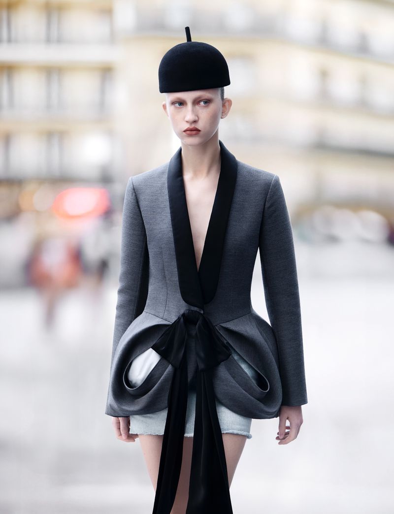 Fashion model in gray jacket and black hat poses on city street with blurred pedestrians and buildings in background, medium shot.