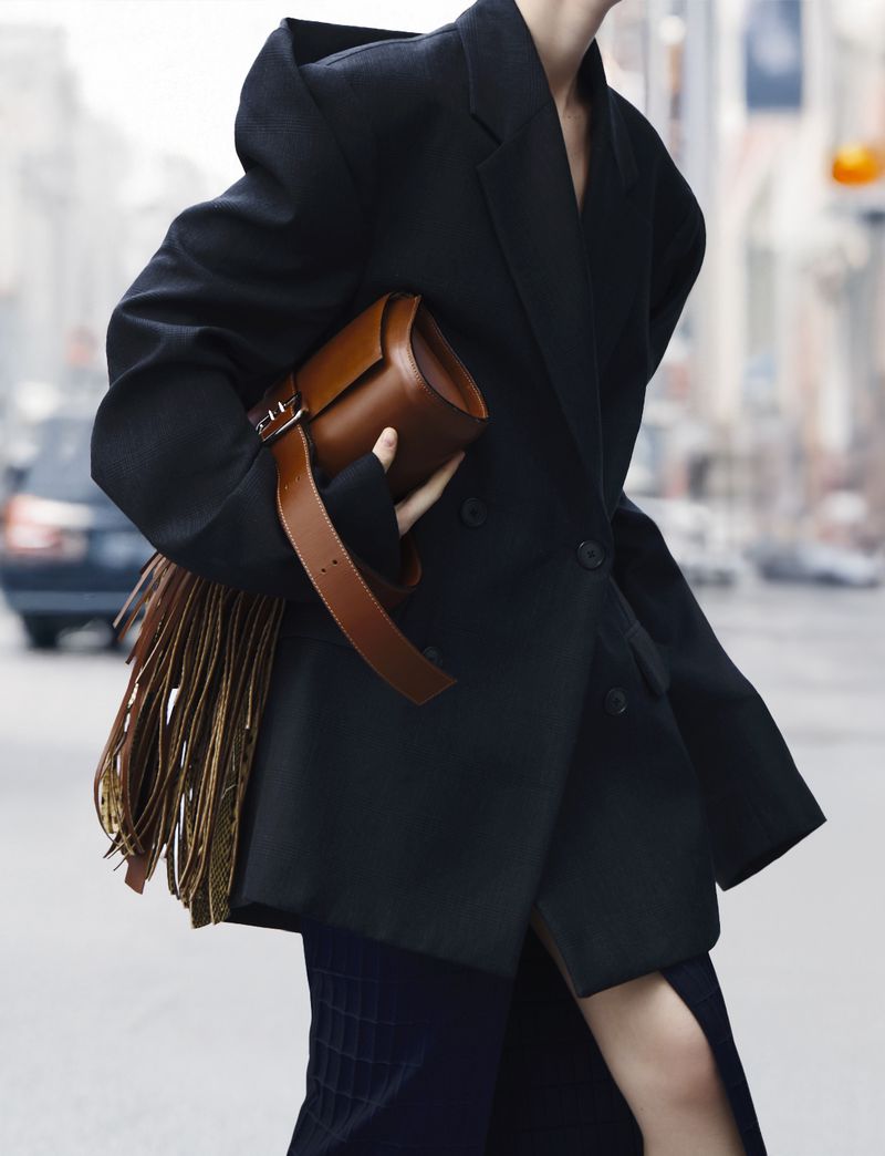 A stylish adult female in an oversized black blazer carries a brown leather fringe bag while walking on a city street.