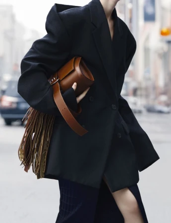A stylish adult female in an oversized black blazer carries a brown leather fringe bag while walking on a city street.
