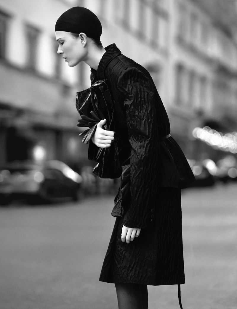 Monochrome fashion editorial image of a model wearing a dark fitted cap and coat holding a large handbag on a city street
