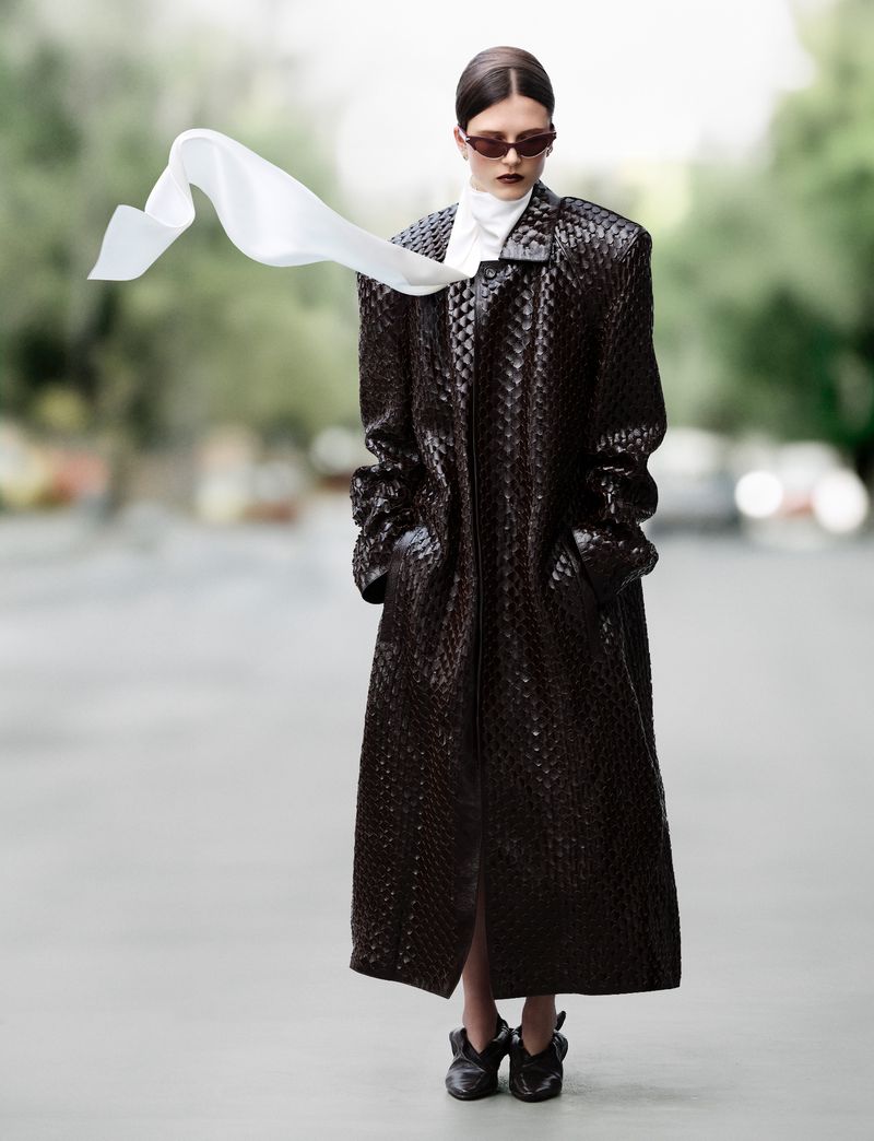 A woman stands on a street wearing a dark brown woven coat, sunglasses, and scarf flowing in the wind, embodying fashion and urban style.