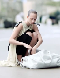 Model crouching outdoors next to a large white leather duffel bag wearing a sleeveless cream top and black pants