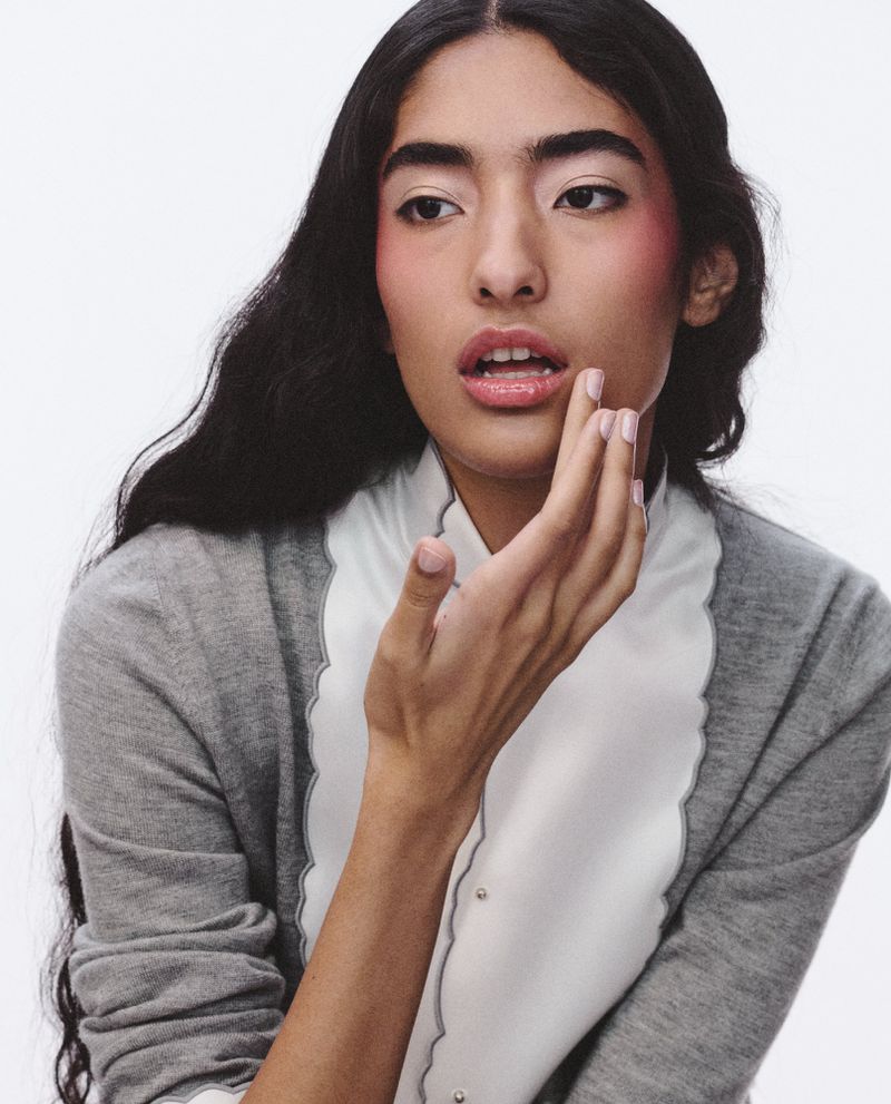 Portrait of a woman with dark hair and fair skin wearing a grey cardigan and white top, posing in a brightly lit photo studio.