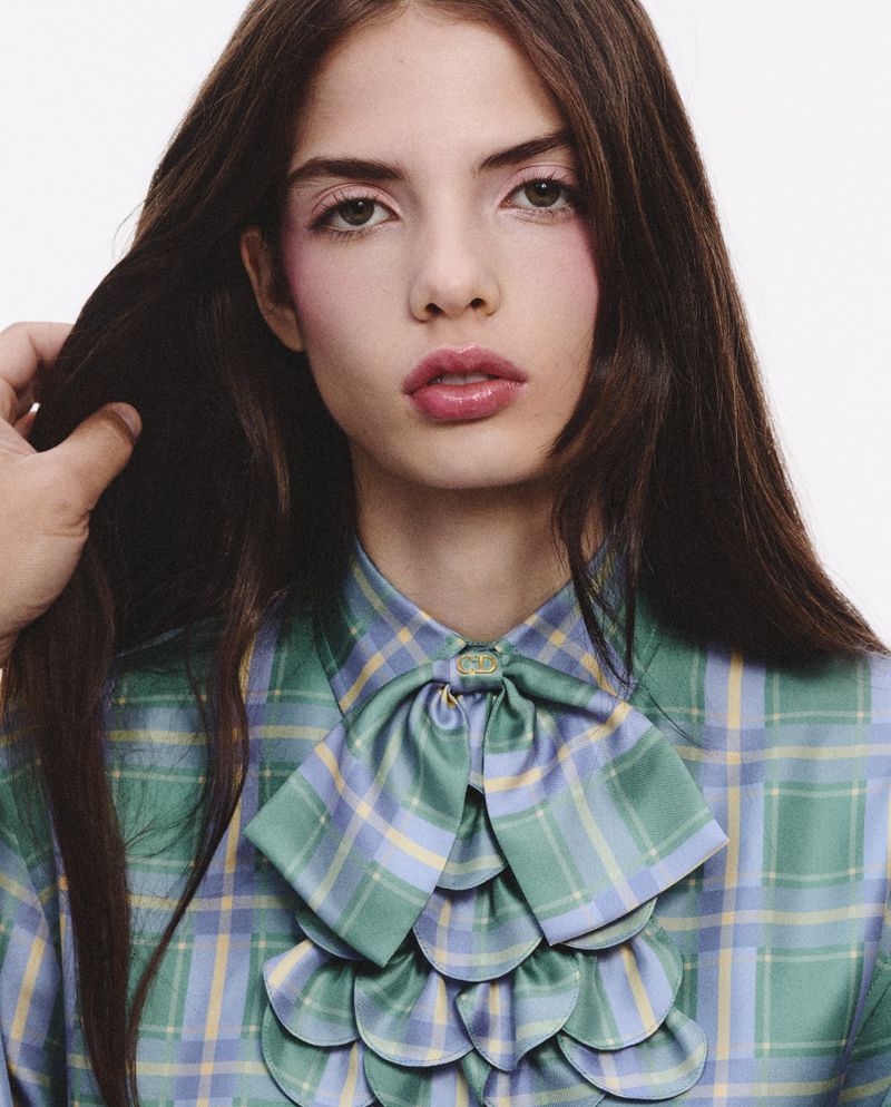 Brunette woman poses in plaid shirt with ruffled collar and an arm adjusts her hair in a studio setting, fashion portrait.