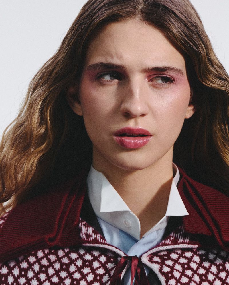 Portrait of a young adult female with wavy brown hair, wearing a red and white knit garment, and vibrant pink makeup.