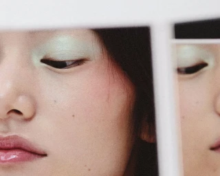 Extreme close-up portrait of a model with pale green eyeshadow and glossy pink lips reflected in a mirror.
