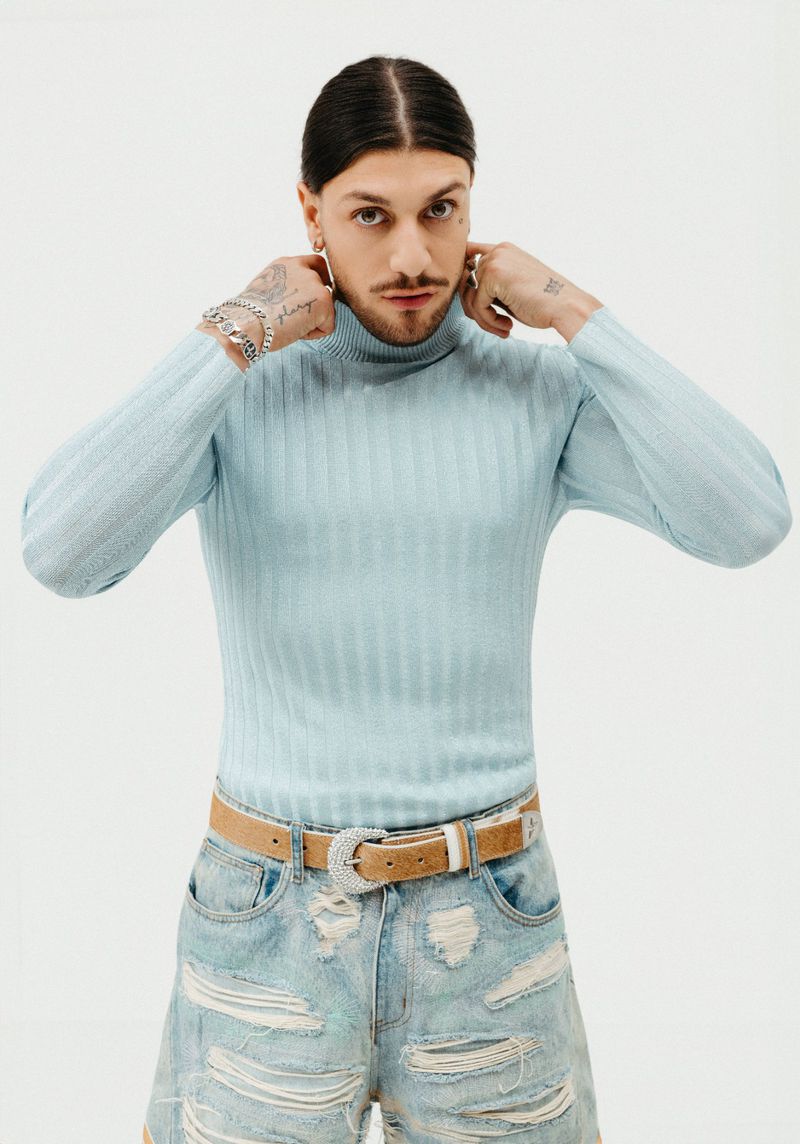 Portrait of adult male with dark hair and mustache wearing a ribbed light blue turtleneck sweater, tan belt, and distressed jean shorts on white.