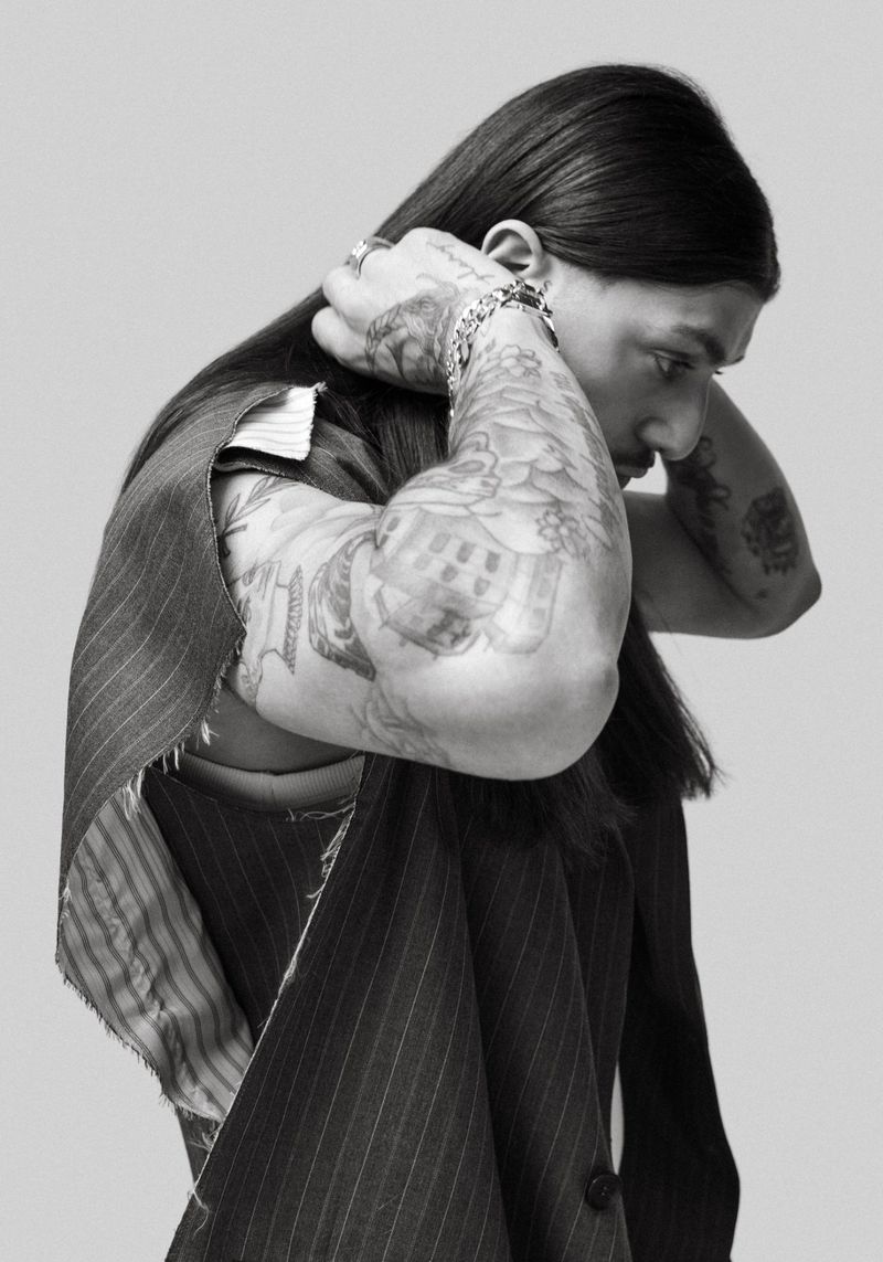 Monochrome portrait of a tattooed adult male with long dark hair, wearing a pinstripe vest, posing in a photo studio.