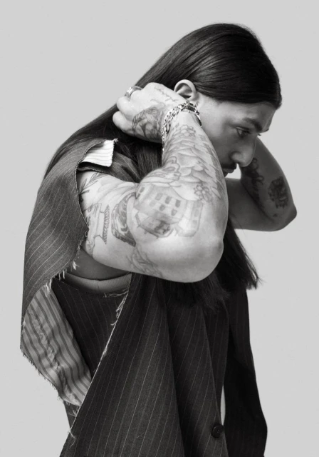 Monochrome portrait of a tattooed adult male with long dark hair, wearing a pinstripe vest, posing in a photo studio.