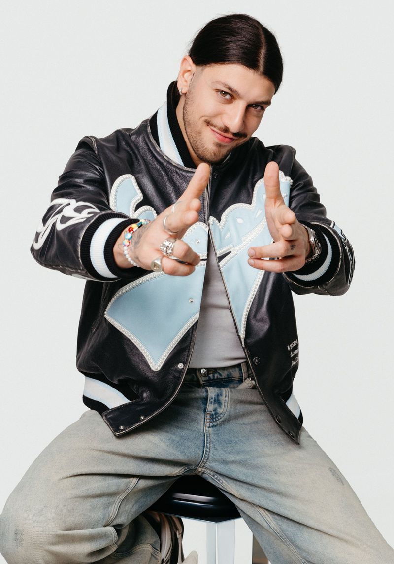 Portrait of a young adult male seated, wearing a black-and-blue jacket and jeans, with rings and bracelets, smiling, on a white background.